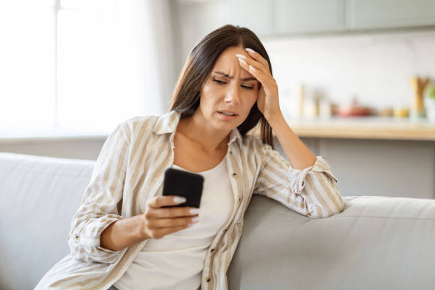 istock Frustrated young woman looking at smartphone while sitting on couch at home 2000547203