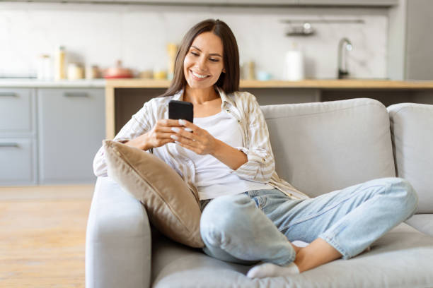 istock Cheerful young woman texting on her smartphone, sitting cross-legged on comfortable couch 2000547210