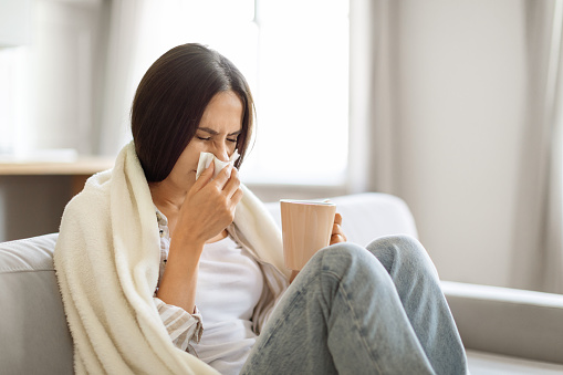 Young woman feeling sick at home, blowing her nose with a tissue and holding mug with tea, ill millennial female wrapped in blanket sitting on couch in living room interior, suffering seasonal flu