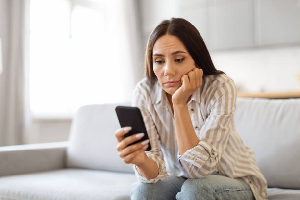 istock Upset Young Woman Sitting On Couch And Looking At Smartphone Screen 2000547765