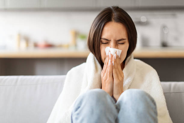 istock Sick Young Woman Blowing Nose In Paper Tissue While Sitting On Couch 2000547800