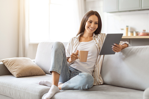 Smiling Young Woman Using Digital Tablet And Drinking Coffee While Relaxing On Couch At Home, Happy Millennial Female Reading E-Book Or Browsing Internet, Resting In Cozy Living Room Interior