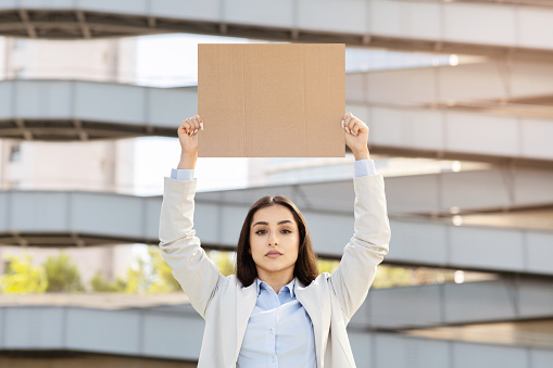 Confident serious young woman holding up blank cardboard sign overhead against an urban backdrop, signifying protest or advocacy. Activism or expression, problems at work, business