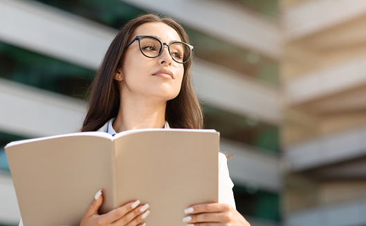 Serious calm confident millennial woman in glasses enjoy work, study, read book in city office outdoors, look at copy space, close up. Business planning and lifestyle, ad and offer, building