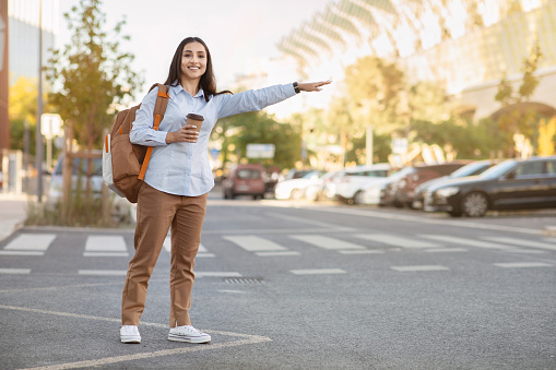 Cheerful pretty young caucasian woman tourist with backpack, cup of coffee takeaway, stop taxi with hand sign in city outdoors. Lady enjoy trip, travel, lifestyle, ad and offer