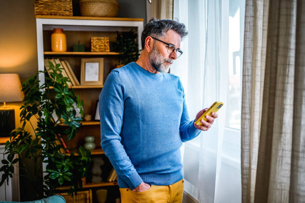 Mature man using phone at home Mature man standing by the window at home and using a mobile phone worried man standing stock pictures, royalty-free photos & images