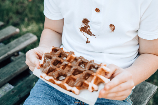 Close up of a child eating Belgian waffles with sticky chocolate hands. Spoiled clothes. outdoors