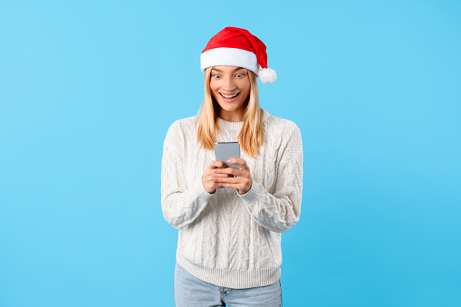 Delighted young female in cozy sweater and Christmas hat engaging with her smartphone, showcasing joyful reaction on plain blue background