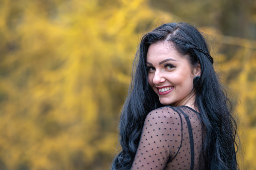 Portrait of a young brunette girl with long black hair  and in a black dress in the autumn park