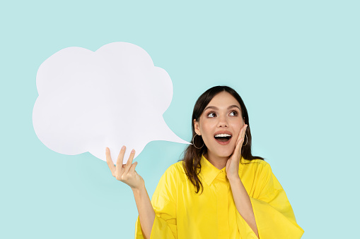 Comment. Portrait of emotional young lady having idea or thought, holding blank speech bubble cloud above her head. Free place for your text, blue studio background, copy empty space
