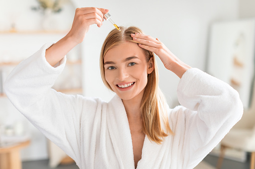 Joyful woman in white robe treats her hair roots for better growth applying nourishing serum or oil via dropper, caring for herself in modern bathroom, smiling to camera