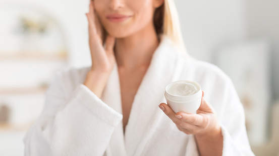 Cropped view of young blonde lady holding jar of moisturizer, enjoying routine of cleansing and hydration with skincare product, standing in bathroom at home, wearing white bathrobe. Panorama