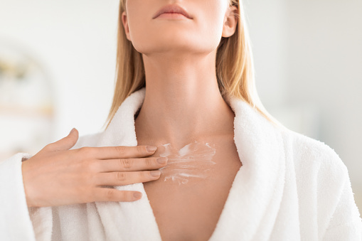 Cropped shot of young blonde woman in bathrobe gently applies moisturizer on her neck, focusing on skincare in her home bathroom. Beauty routine and care to maintain smooth, healthy skin