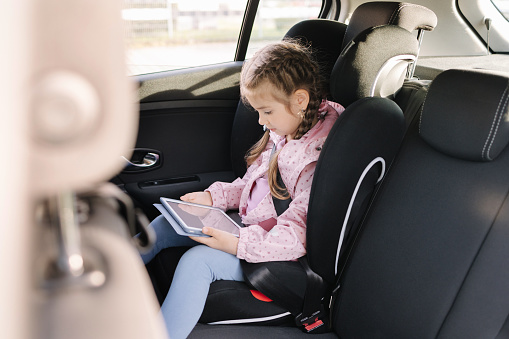 Cute little girl use tablet in car seat on the way to the kindergarten. High quality photo