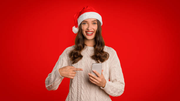 istock Smiling woman in Santa hat pointing at cell phone, studio 2000639196