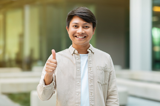 Positive young asian entrepreneur showing thumb up and smiling at camera, posing over office building at downtown. New career opportunities, business, entrepreneurship