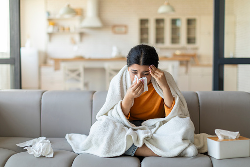 Ill indian woman blowing her nose with tissue at home, sick eastern female sitting wrapped in blanket on couch in living room, feeling unwell, showing symptoms of cold and seasonal flu, copy space