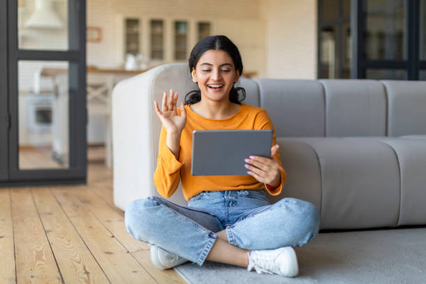 istock Indian woman sitting on floor, waving hello during video call on tablet 2000663507