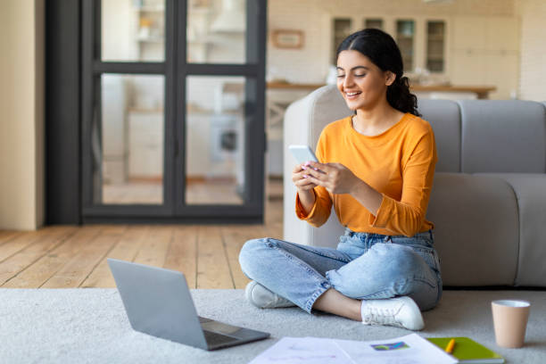 smiling indian woman sitting cross-legged on floor and using smartphone and laptop - multi tasking asian and indian ethnicities asian ethnicity lifestyles imagens e fotografias de stock