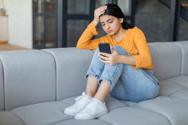 istock Worried young indian woman with smartphone in hands sitting on sofa 2000663533