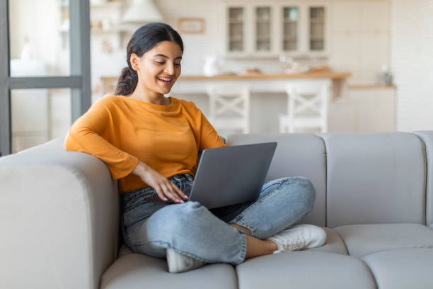 istock Smiling young indian woman using her laptop while sitting cross-legged on couch 2000664054