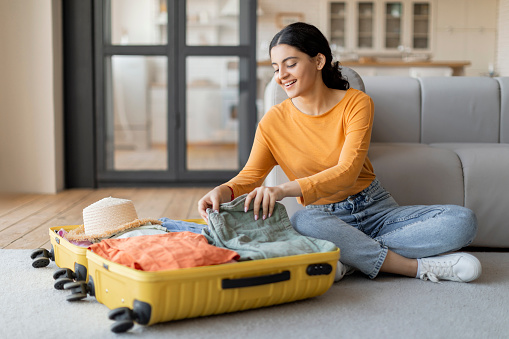 Smiling young indian woman sitting on the floor, contentedly packing clothes into her open yellow suitcase, happy eastern female getting ready for trip, preparing luggage for journey, copy space