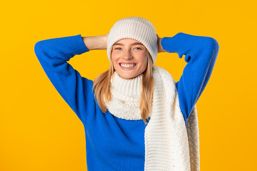 Cheerful woman in blue sweater touches her white knitted hat, exuding happiness and warmth, against yellow background, ready for a cold day