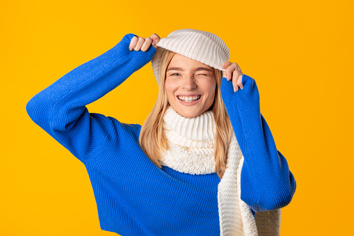 Cheerful woman in blue knit sweater smiling and winking, playfully adjusts her white knitted hat, standing against yellow background, emanating warmth and joy