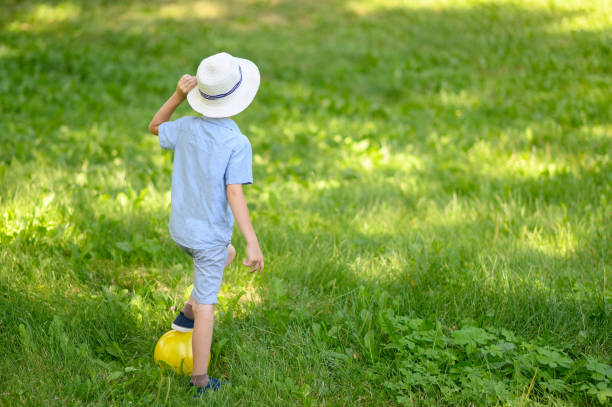 A boy in a hat stands on the grass on a sunny summer day with a yellow ball, view from the back.The concept of vacations, holidays in nature active sports.Copy space.