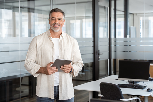 Latin Hispanic mature adult professional business man looking at camera and smiling. Indian senior businessman CEO holding digital tablet using fintech tab application standing inside company office.