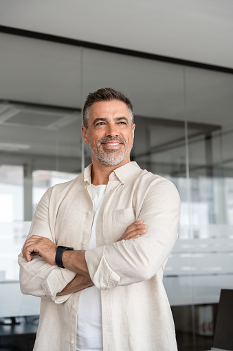 Vertical portrait handsome hispanic senior business man with crossed arms confident smiling. Indian or latin confident mature good looking middle age leader male businessman on blur office background.
