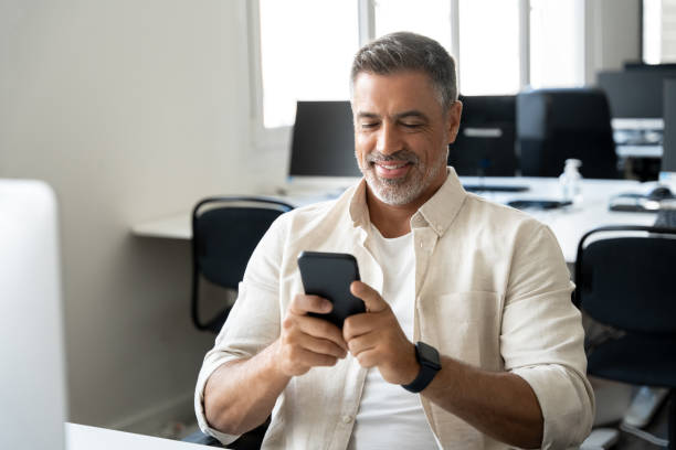 istock Middle aged Hispanic business manager ceo using cell phone mobile app, laptop. Smiling Latin or Indian mature man businessman holding smartphone sit in office working online on gadget with copy space. 2000672150