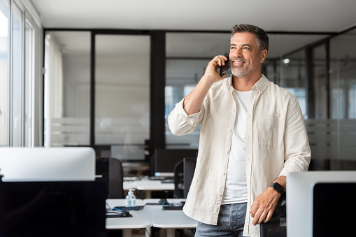 Middle aged Latin or Indian businessman having call on smartphone with business partners or clients. Smiling mature Hispanic man in relaxing pose talking by mobile cellphone at work in modern office.