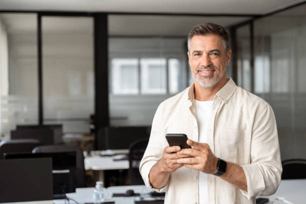 istock Smiling at camera mature Latin businessman holding smartphone in office. Middle aged manager using cell phone mobile app. Digital technology application and solutions for business success development. 2000672182