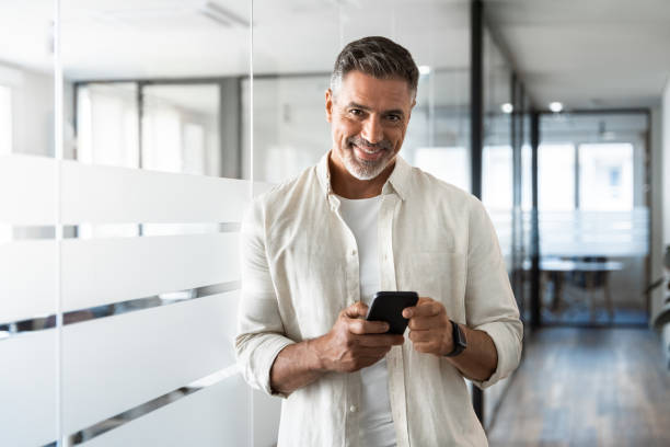 istock Smiling mature Latin or Indian businessman holding smartphone, using cellphone mobile app. Middle aged hispanic employee standing with phone and looking at camera on office background with copy space. 2000672184