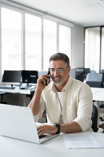 Middle aged Latin or Indian businessman having call on smartphone with business partners or clients. Smiling mature Hispanic man sitting at table talking by mobile cellphone in office, vertical.