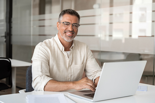 Portrait of mature Indian or Latin business man ceo trader using laptop computer, typing, working in modern office. Middle-age Hispanic smiling handsome businessman entrepreneur looking at camera.