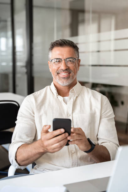 istock Vertical portrait of middle aged Hispanic business manager using cell phone mobile app. Latin or Indian mature man businessman holding smartphone sit in office work online on gadget, smiling at camera 2000672704
