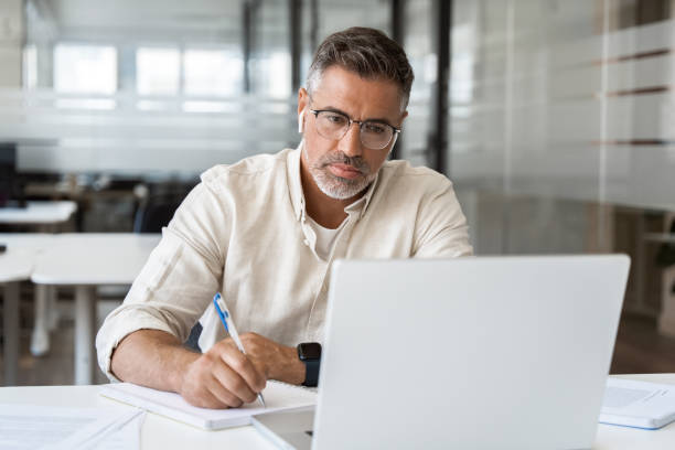 istock Focused middle-age Hispanic man using laptop computer for business studying, watch online virtual webinar training meeting, video call, make notes. Mature Latin businessman work in office, copy space. 2000672711
