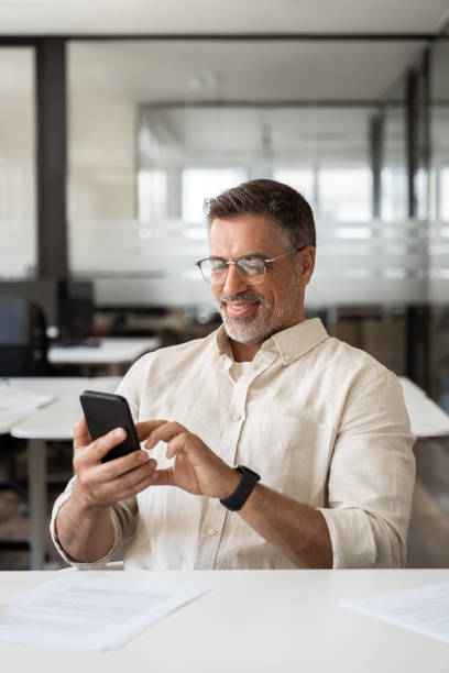 istock Vertical portrait of middle aged Hispanic business manager using cell phone mobile app. Smiling happy Latin or Indian mature man businessman holding smartphone sit in office working online on gadget. 2000672714