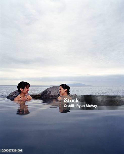 Two Young Women Soaking In Hot Spring Sea In Background Stock Photo - Download Image Now