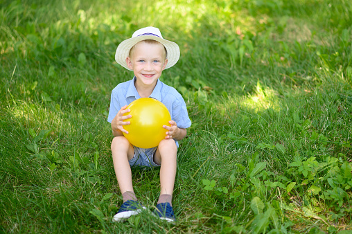 A happy boy in a hat sits on a sunny summer day on the grass and holds a yellow ball in his hands. The concept of vacations, holidays in nature active sports.