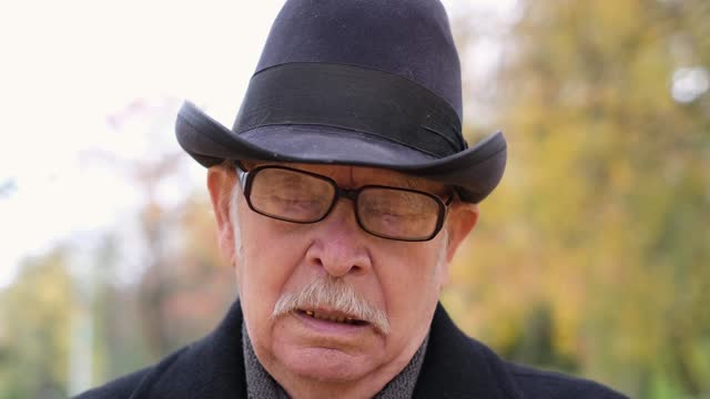 Portrait of a grandfather in a hat who adjusts his glasses while standing in an autumn park
