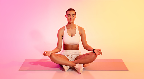 Serene latin young woman with a short haircut in sportswear meditating in lotus position on a yoga mat, eyes closed, against a peaceful pink and yellow gradient background