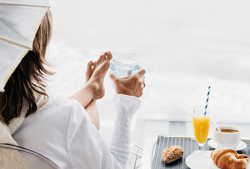 a woman sitting on a terrace, having breakfast, holding a glass of water while contemplating the sea. concept of healthy living and relaxation.