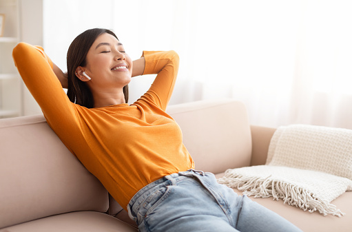 Relaxed smiling millennial asian woman reclining on couch with hands behind her head and closed eyes, using wireless earphones, listening to music or favorite podcast at home, copy space