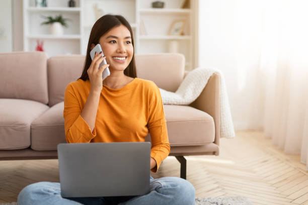 Smiling millennial asian lady using laptop and speaking on smartphone Smiling millennial asian lady using laptop and speaking on smartphone while sitting on floor in living room, looking at free space. Lovely young woman having phone conversation, working on pc project manager remote stock pictures, royalty-free photos & images