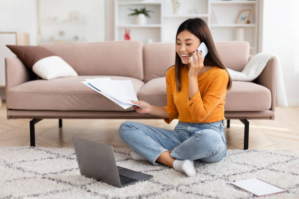 Asian woman freelancer working from home, talking on cell phone Cheerful millennial Asian woman freelancer working from home, sitting on carpet floor next to couch, holding papers, using laptop computer and talking on cell phone. Remote work, freelance project manager remote stock pictures, royalty-free photos & images