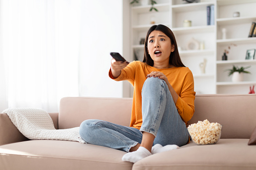 Shocked millennial asian woman watching TV at home, sitting on couch in living room interior, holding remote control from television, eating popcorn, watching horror, thriller, news, copy space