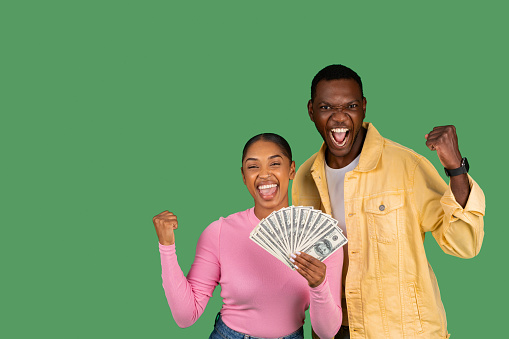 Emotional happy lucky young african american couple man and woman celebrating success together, showing money cash dollars, clenching fists and exclaiming, isolated on green studio background